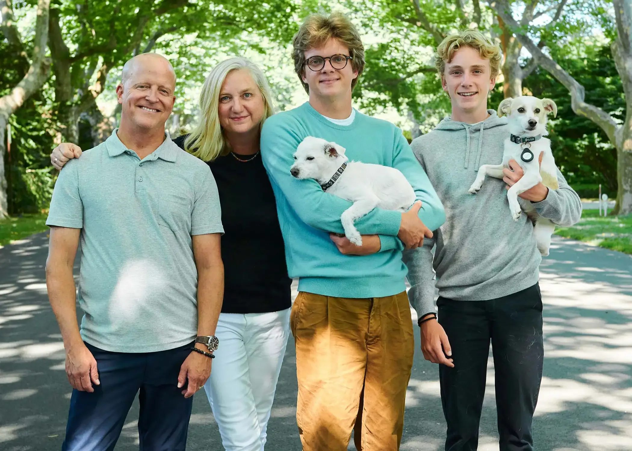 Family of five standing on a street in a park, photographed with two dogs, surrounded by green trees.