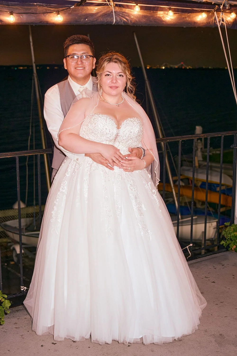 A newlywed couple stands close together on a boat at night. The bride wears a white lace wedding dress and veil, and the groom is in a beige vest and white shirt with glasses. The background features water and a dark sky, illuminated by string lights