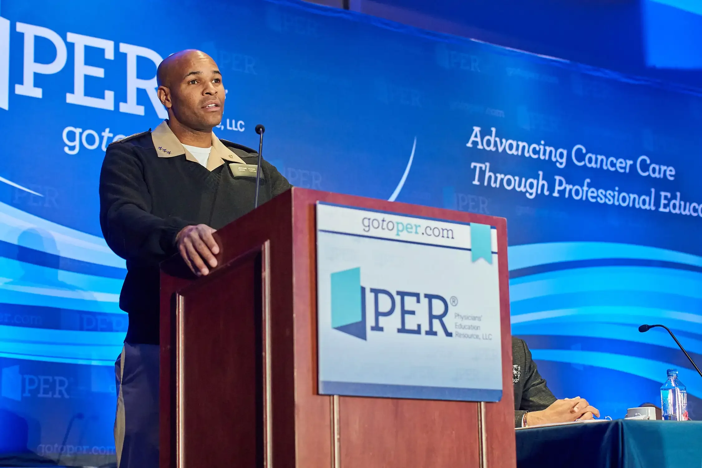 A man in military uniform speaking at a podium during a conference. The background displays the logo and slogan of the conference, 'Advancing Cancer Care Through Professional Education,'