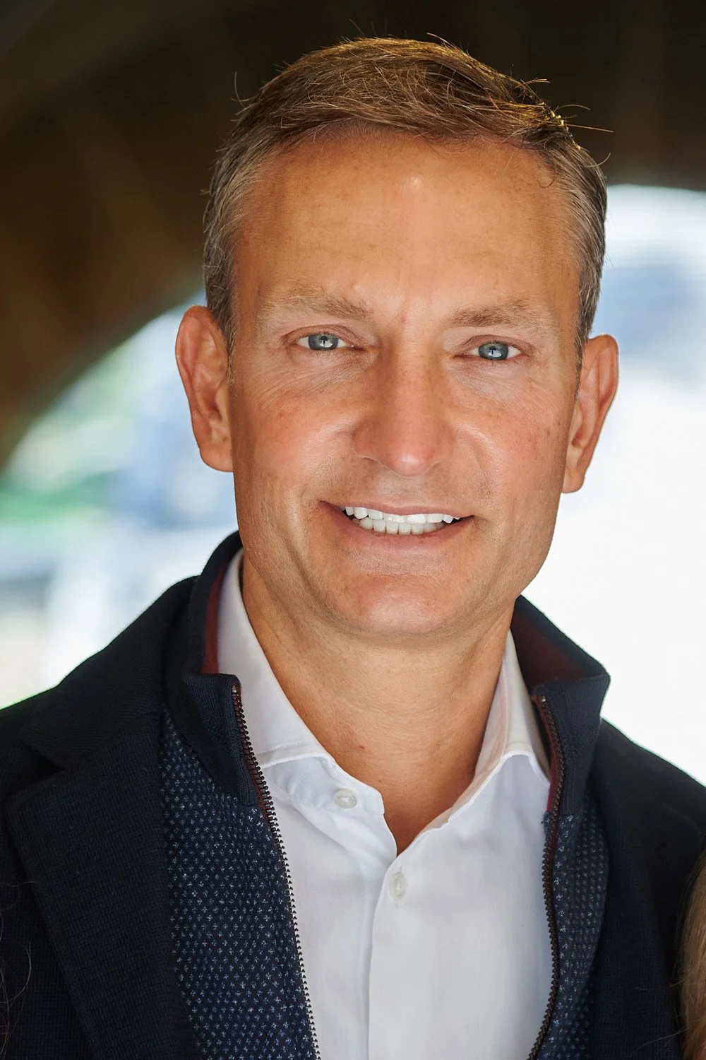 A smiling man with blue eyes, short brown hair, wearing a white collared shirt and black jacket, outside with blurred background.