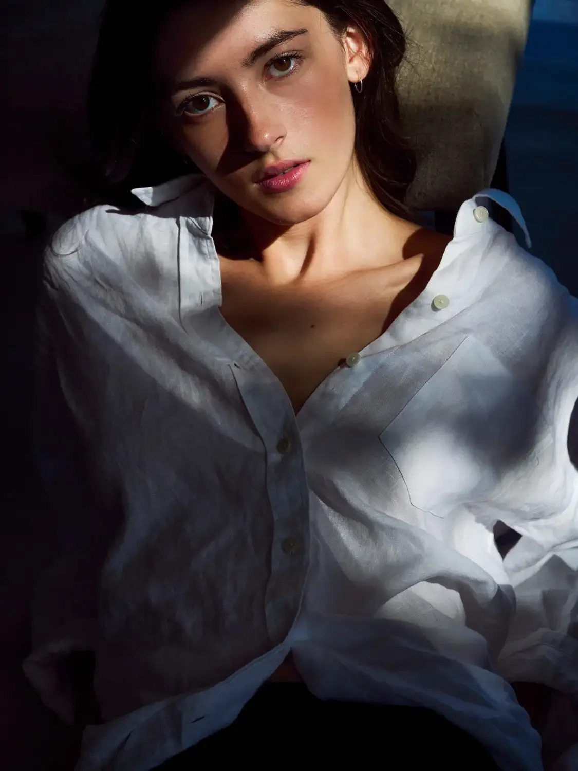 A young woman with dark hair and brown eyes wearing a white button-up shirt, sitting in the sunlight, looking directly at the camera.