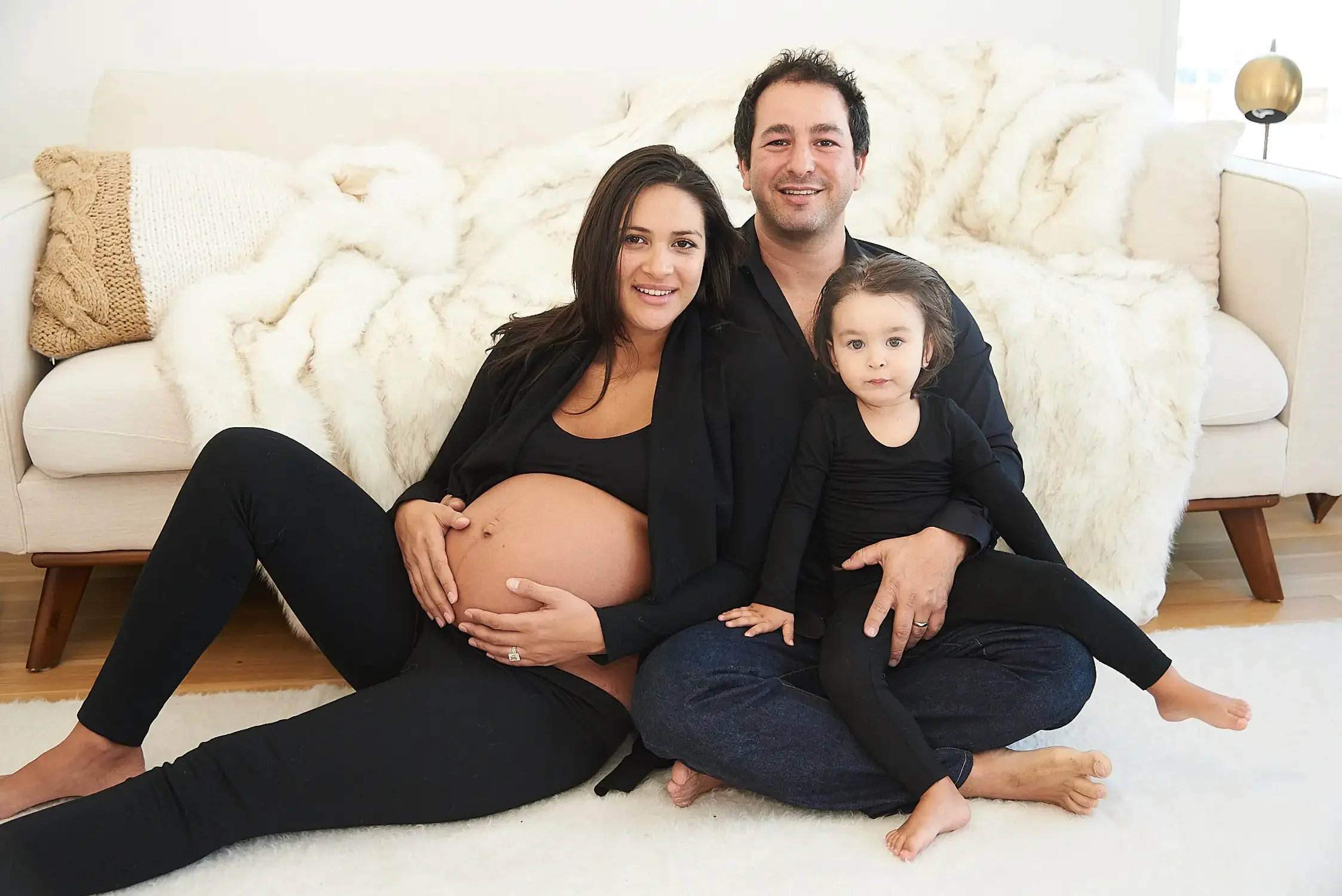 A family of three sitting on a white floor with a white couch in the background. The woman is pregnant and sitting with her legs extended to the side, while the man and young girl sit close, all smiling.