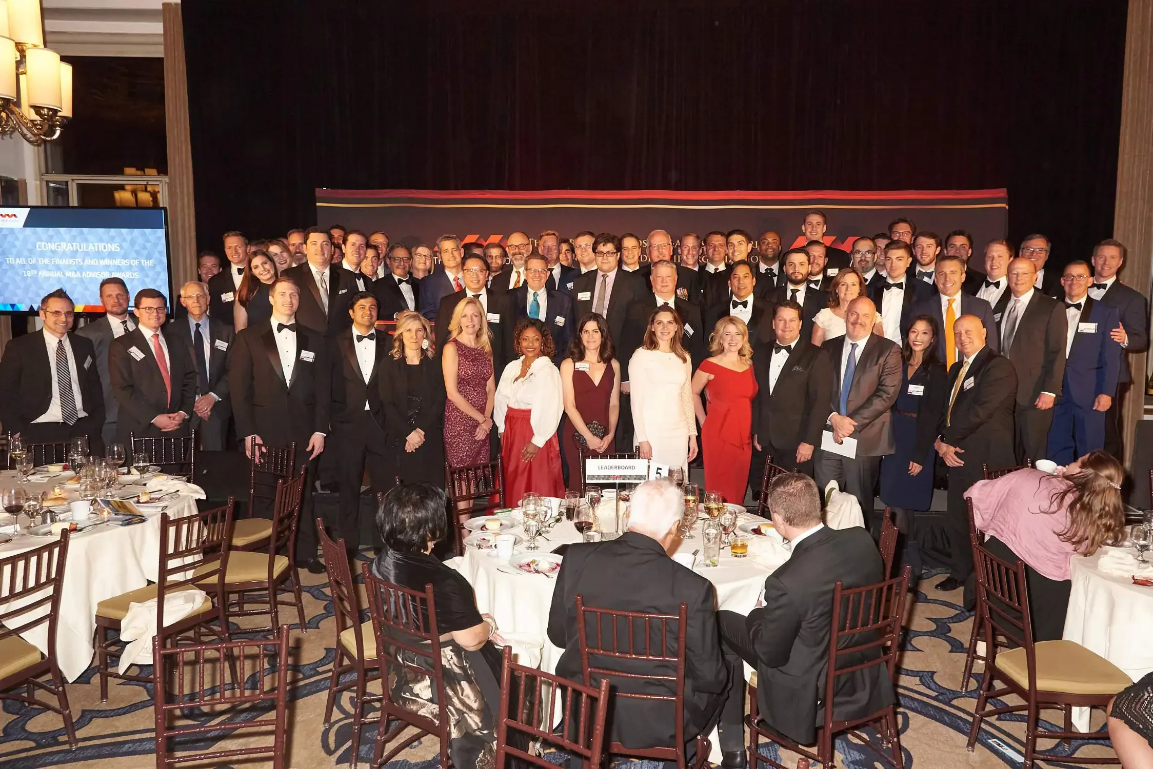 A large group of people dressed in formal business attire, including tuxedos and elegant dresses, gathered on stage for an awards ceremony.