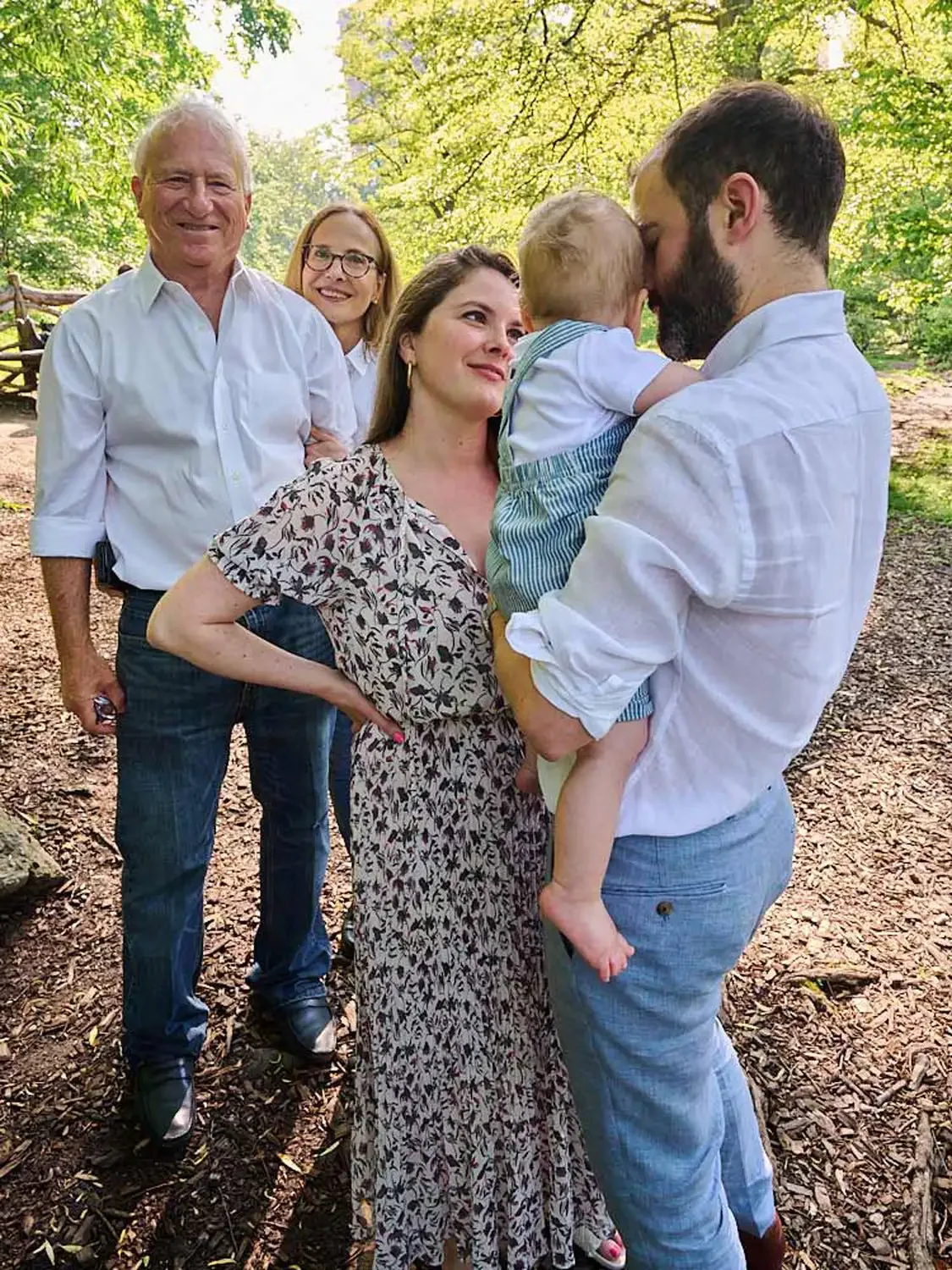 Family of five photographed standing outdoors under trees, with a woman holding a young child, and three adults smiling.