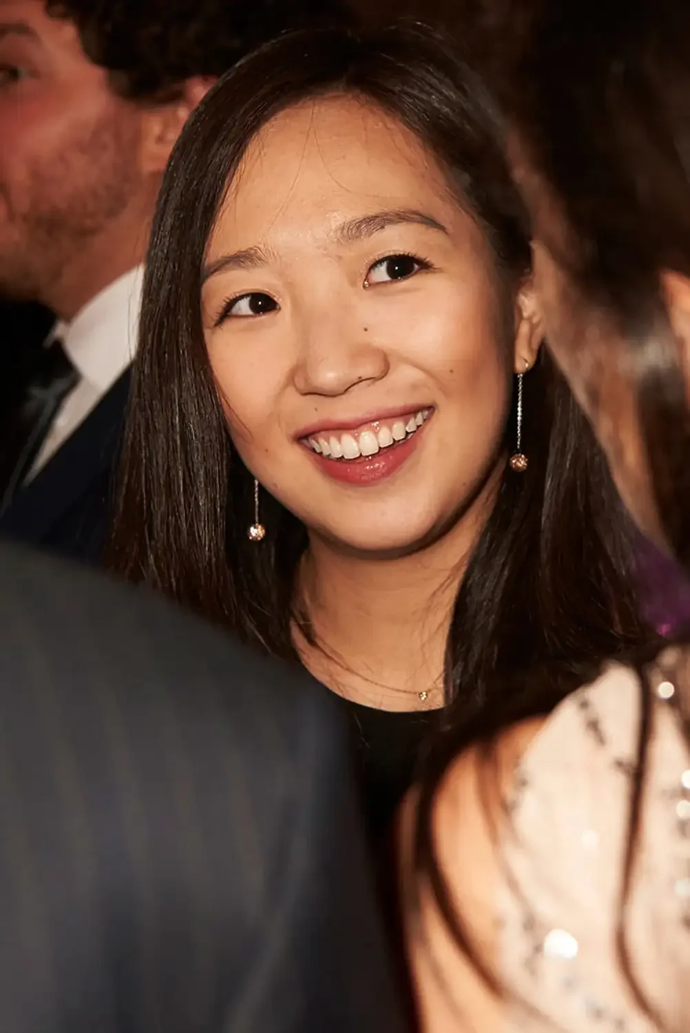 A young stylish Asian woman with shoulder-length hair smiling and looking at someone at a corporate social event, wearing earrings and a black top.