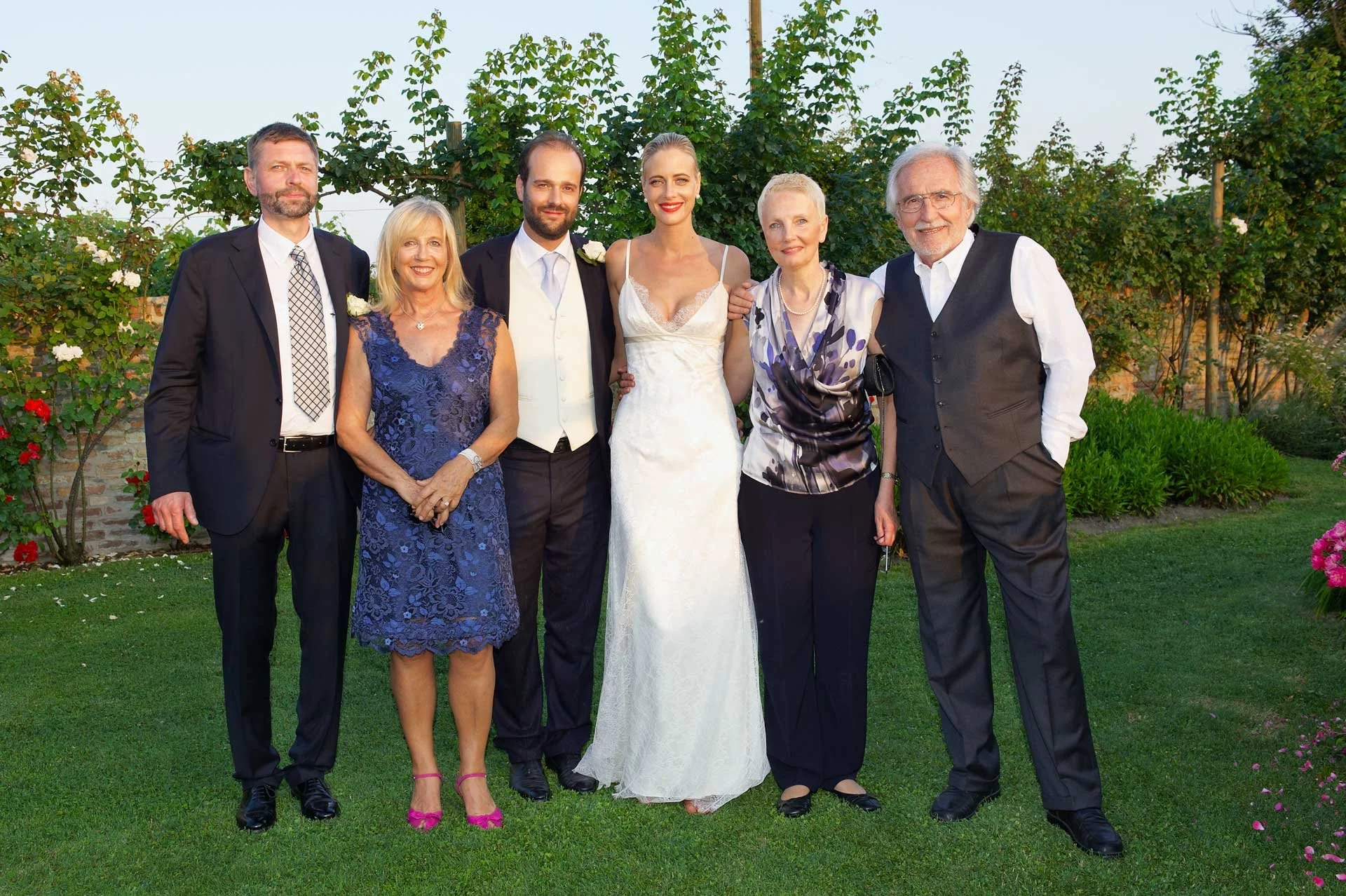 Group of seven people standing outdoors on grass, dressed in formal attire, with a backdrop of green foliage and flowers, during a wedding celebration.