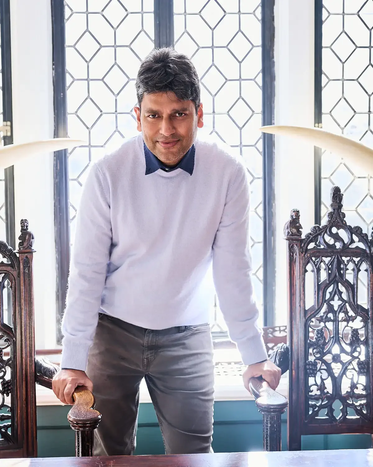 Portrait of a man with dark hair and a light complexion standing in front of a window with geometric latticework, wearing a white sweater over a navy blue shirt.