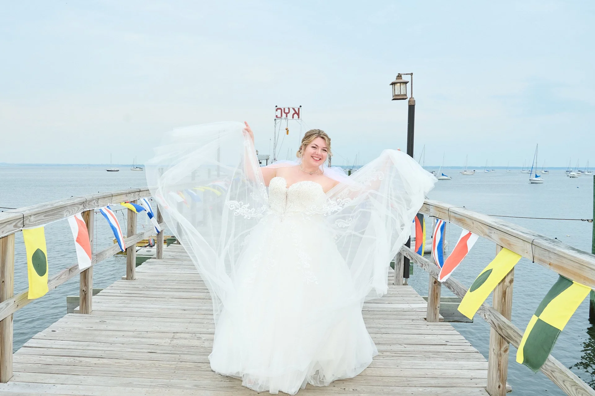 A woman in a wedding dress is standing on a wooden pier by the water, holding the sides of her dress out to show its full length. There are boats in the background, and nautical flags decorate the sides of the pier.