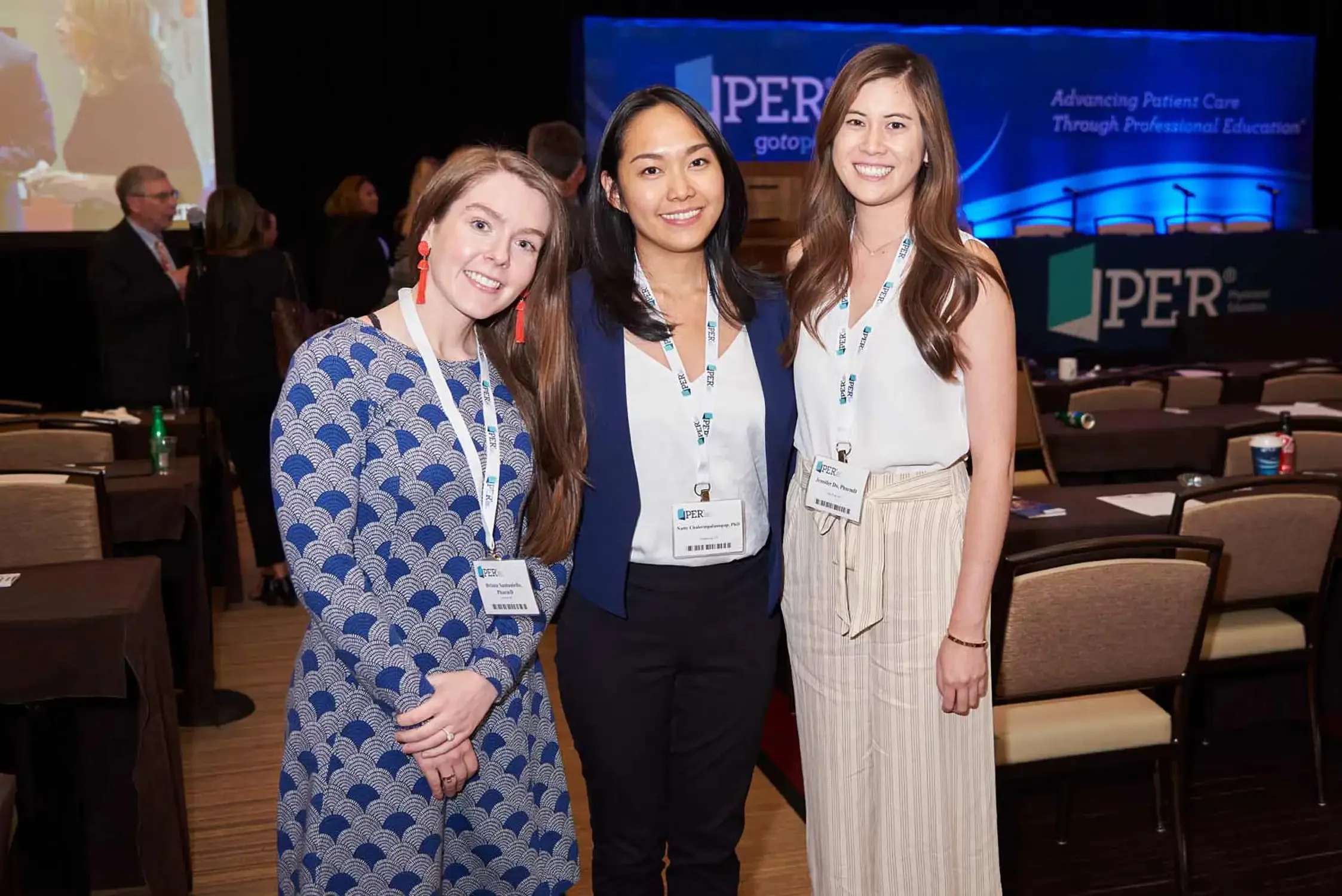 Three women standing together at a conference, smiling, with a conference banner in the background. They are wearing conference badges at a corporate event.