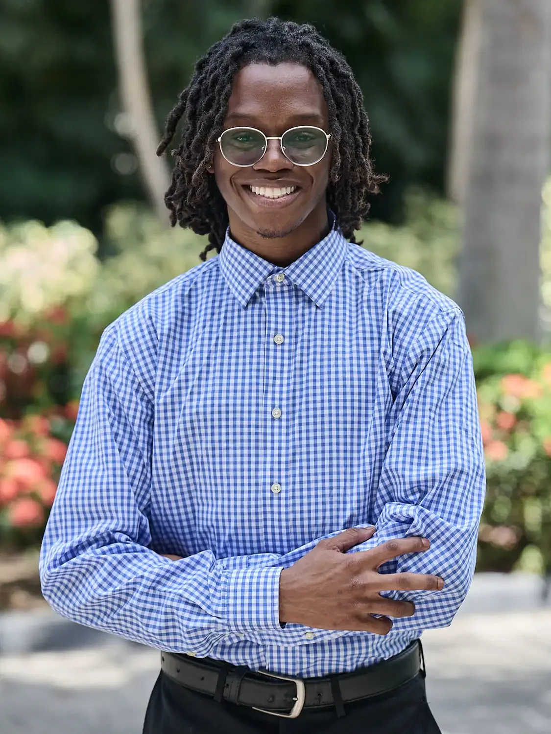 A man wearing glasses and a blue checkered shirt smiles with arms crossed outdoors smiling and friendly photographed in a professional way.