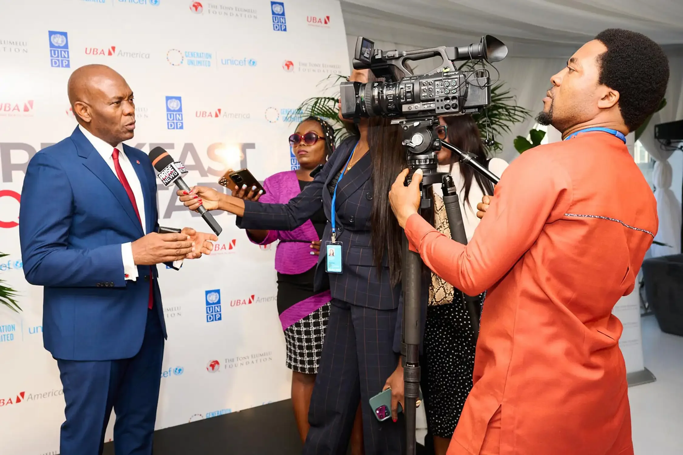 A man in a blue suit and red tie is being interviewed by a woman with long hair, glasses, and a black and white checkered skirt, using a microphone at a corporate event.