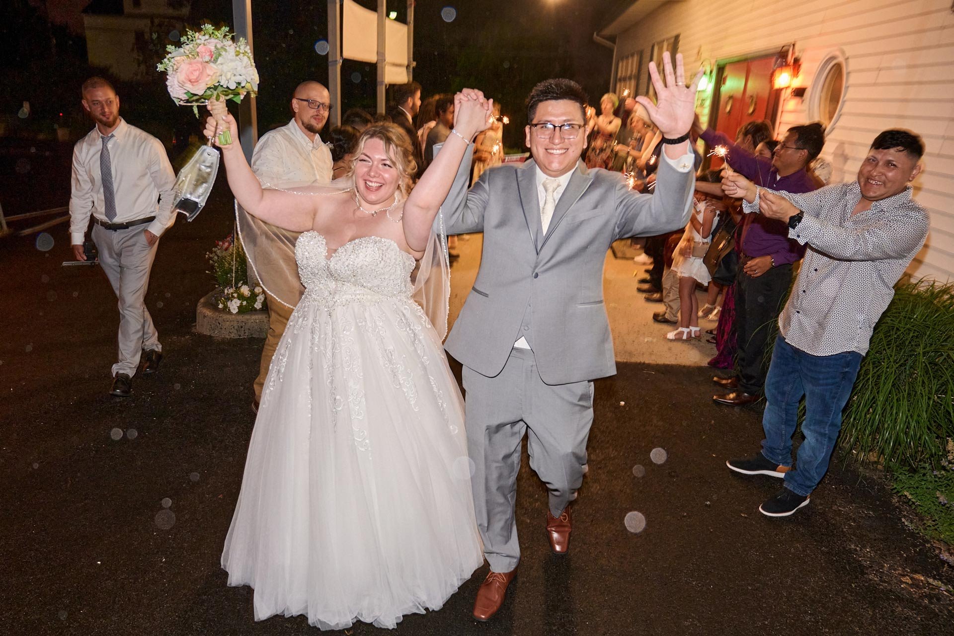 A newlywed couple happily walking together while holding hands, surrounded by friends and family celebrating with sparklers at night.