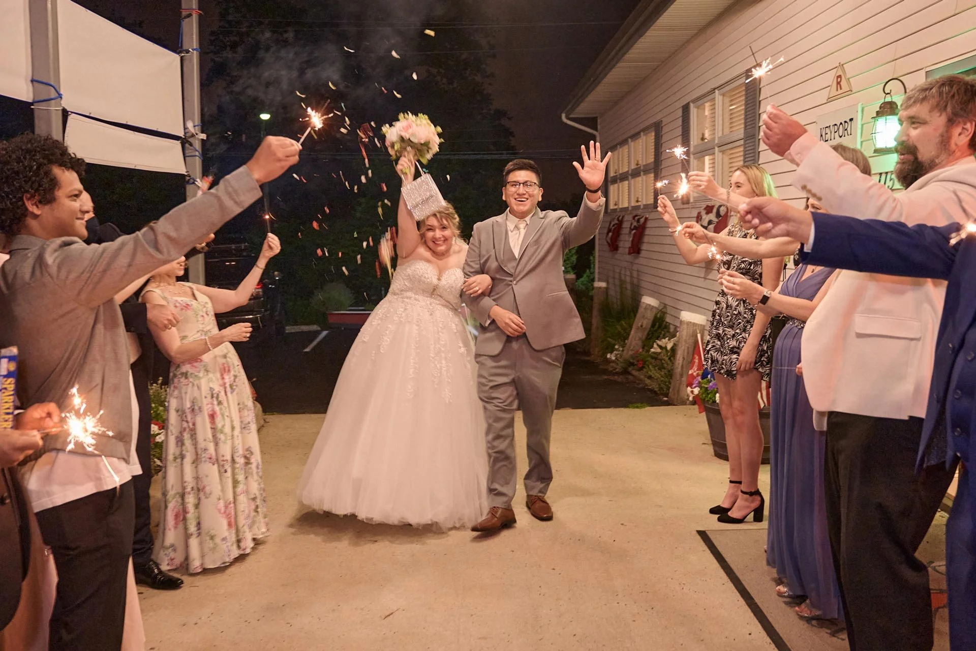 A bride and groom walking arm-in-arm through a sparkler send-off, smiling, with guests holding sparklers on either side at night outside a building decorated for a wedding celebration.