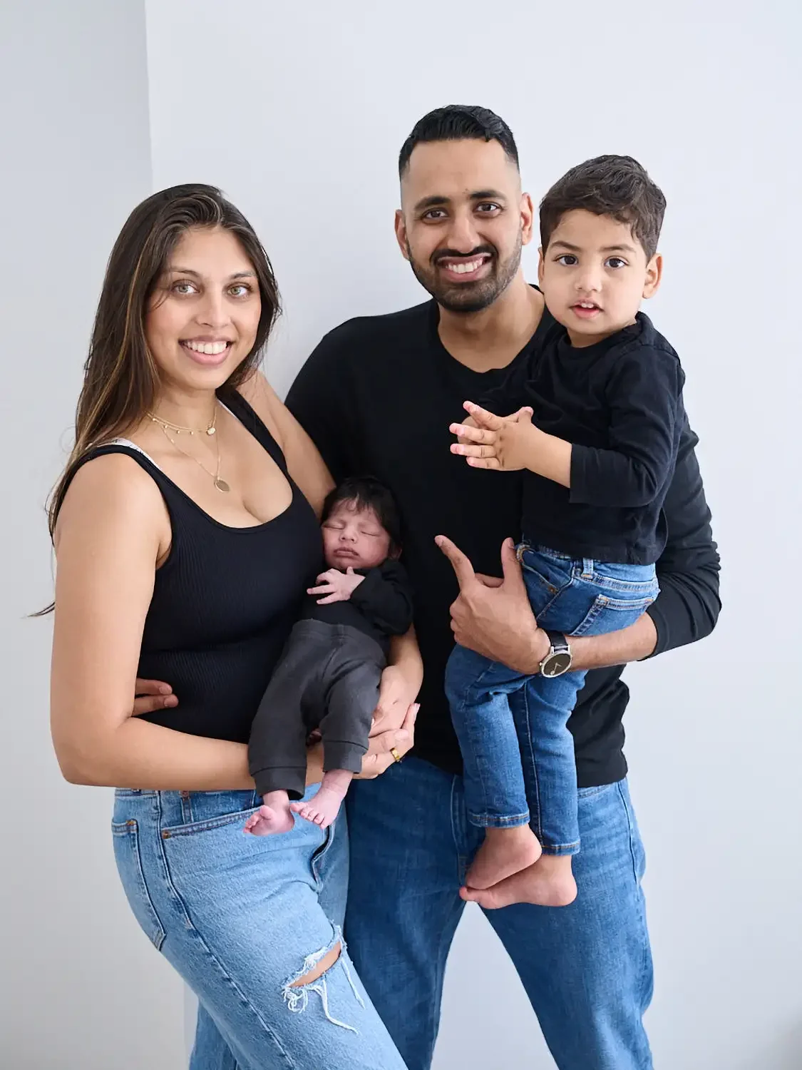 A happy family of four, including a woman, a man, a young boy, and a newborn baby, smiling and posing together against a plain wall.