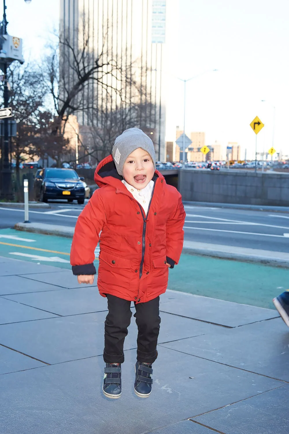 A young boy wearing a red jacket, gray beanie, and black pants is jumping in the air on a city sidewalk during daytime. Cars and tall buildings are visible in the background.