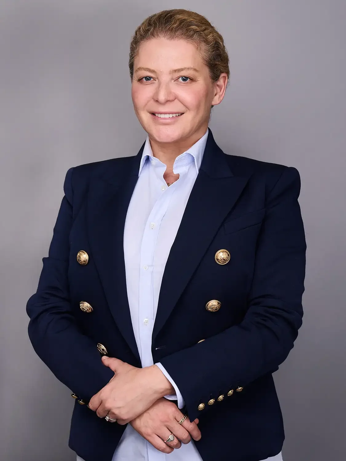 Headshot of a woman in a navy blazer with gold buttons and a white shirt, smiling photographed in a professional photo studio.