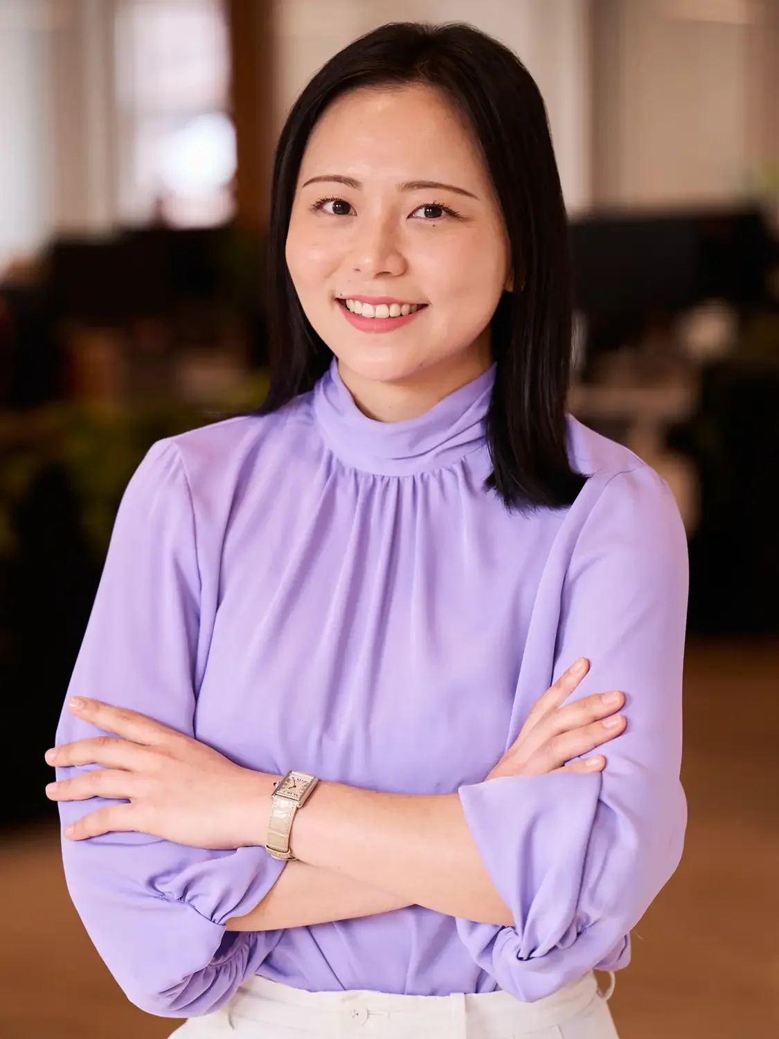 An asian woman with black hair wearing a lavender blouse, smiling with arms crossed, in an office setting reflecting confidence.