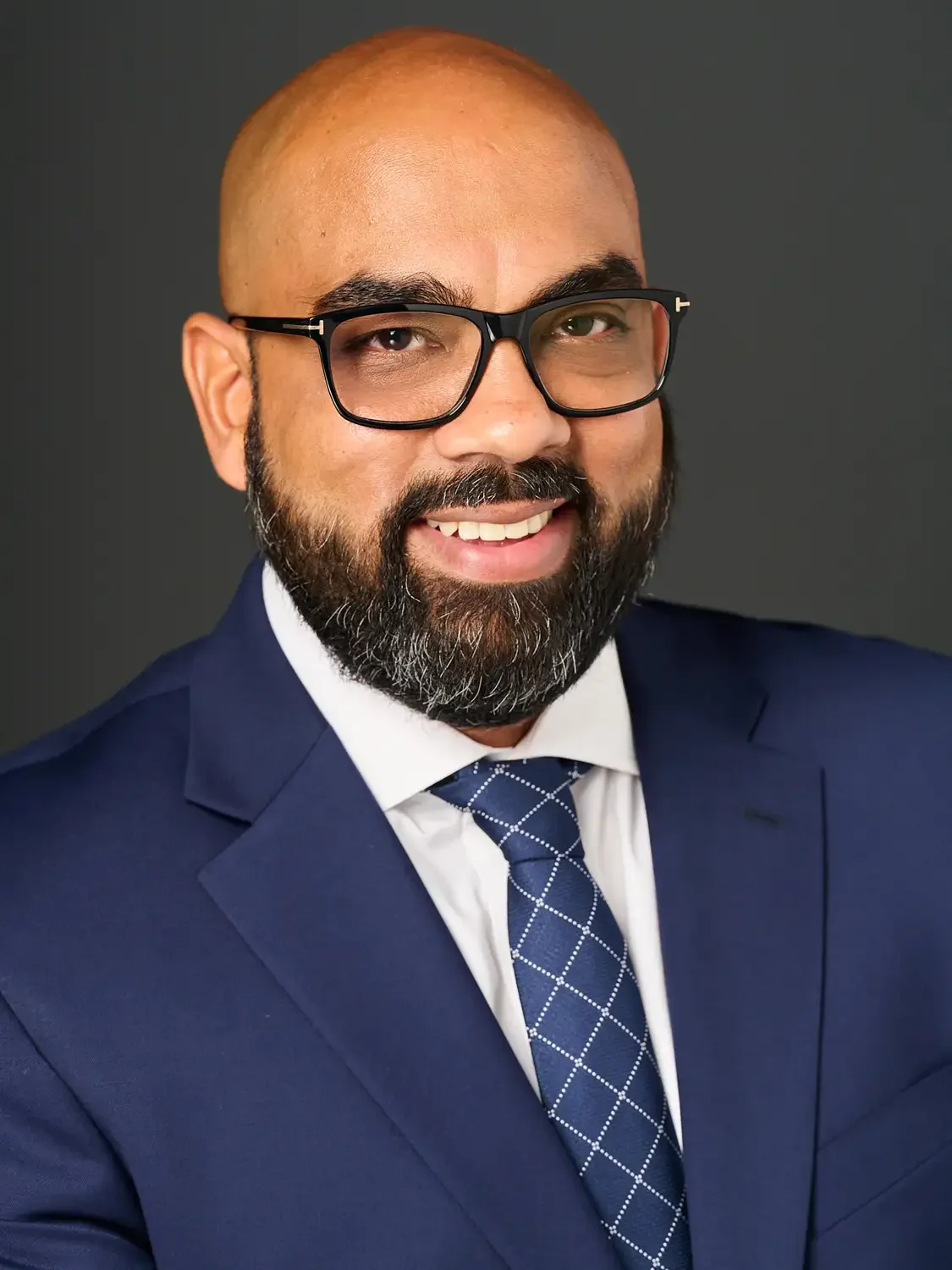 Portrait of a man with a beard and glasses wearing a navy blue suit, white shirt, and patterned tie in a photo studio with dark gray background.