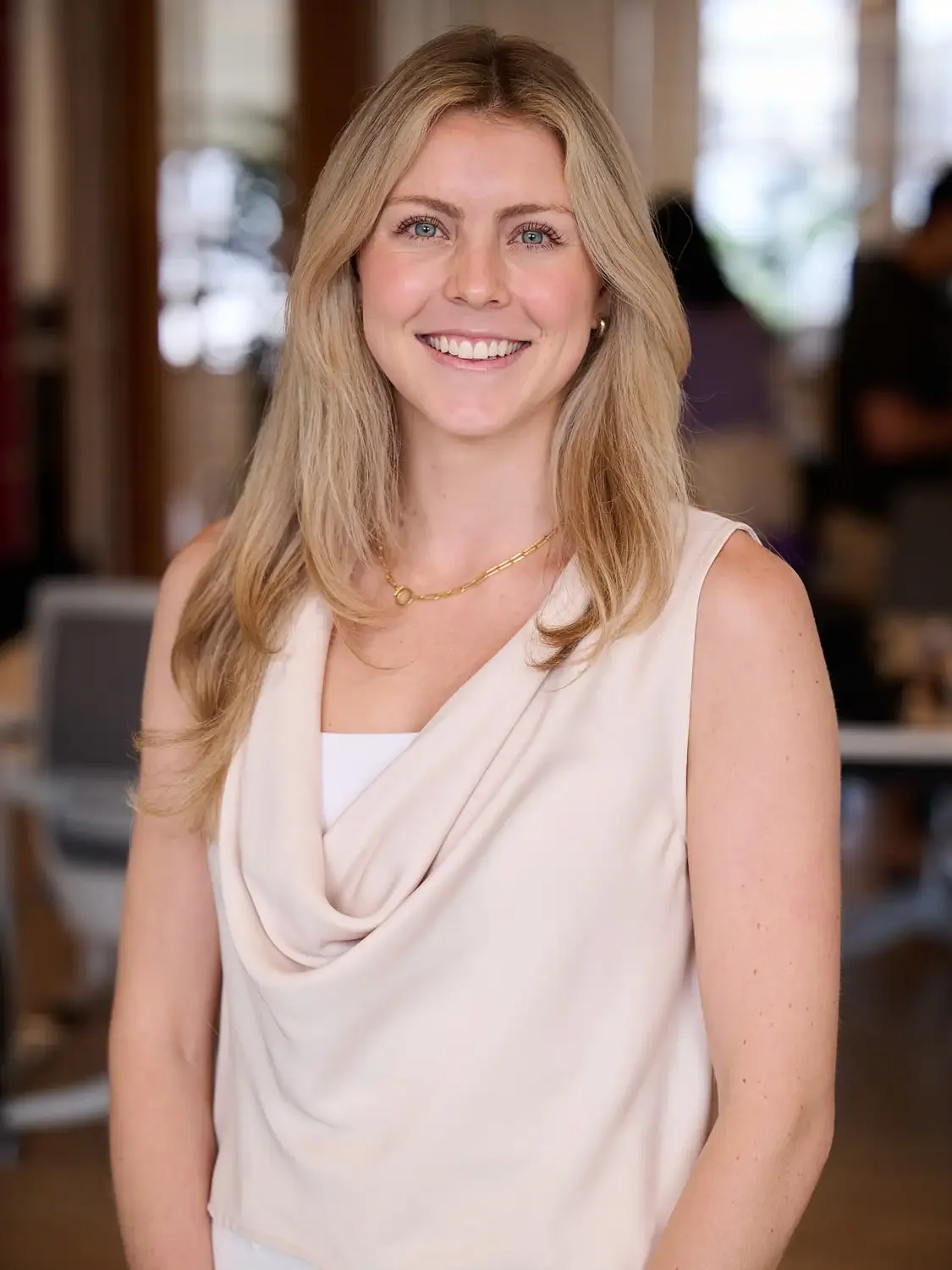 Portrait of a woman with blonde hair, wearing a sleeveless cream-colored top, smiling in an office setting photographed in an elegant lighting.