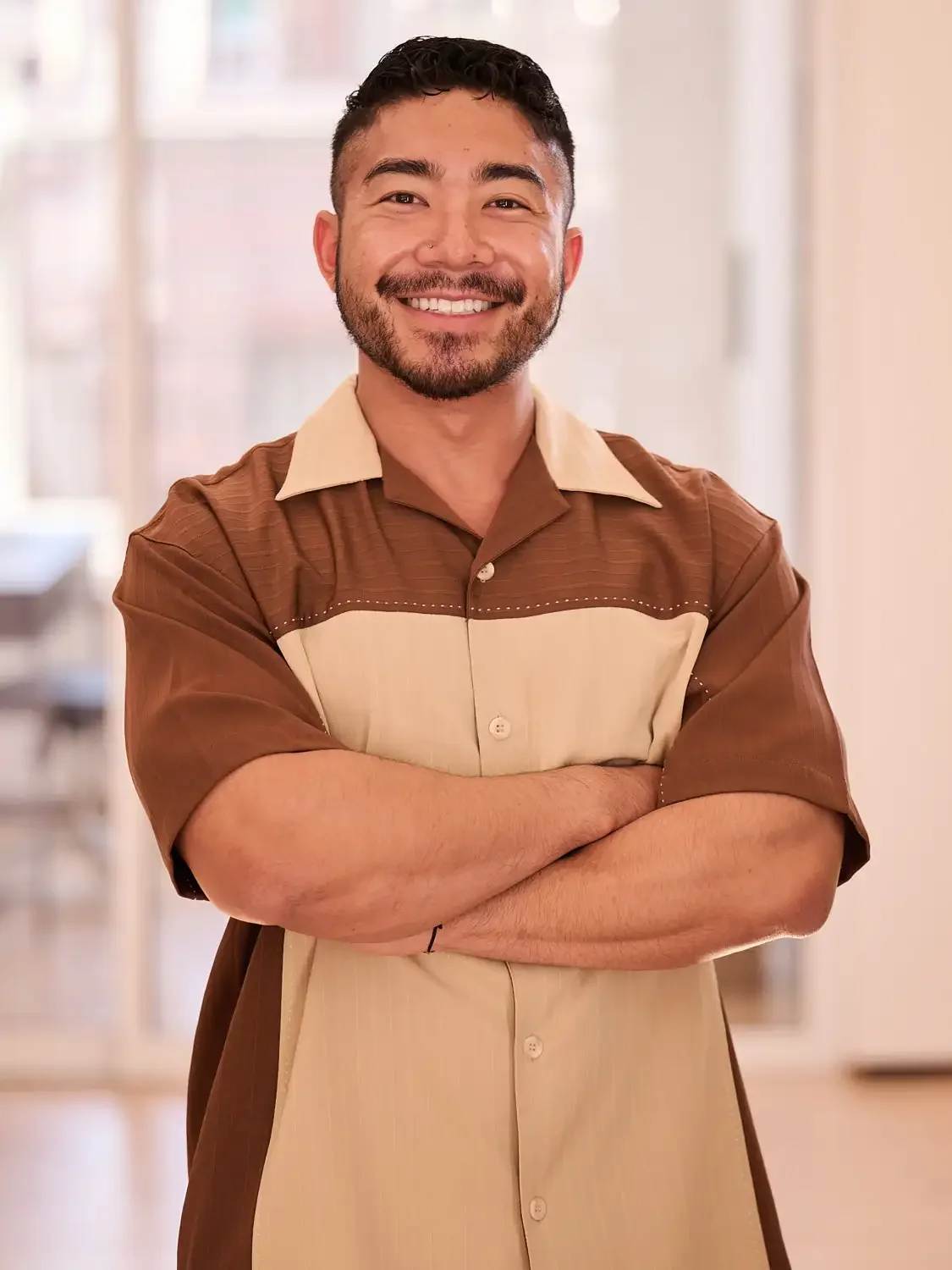 Portrait of a man with dark hair and a beard, smiling, standing with arms crossed in an office.  Professional headshot capturing confident workplace image.