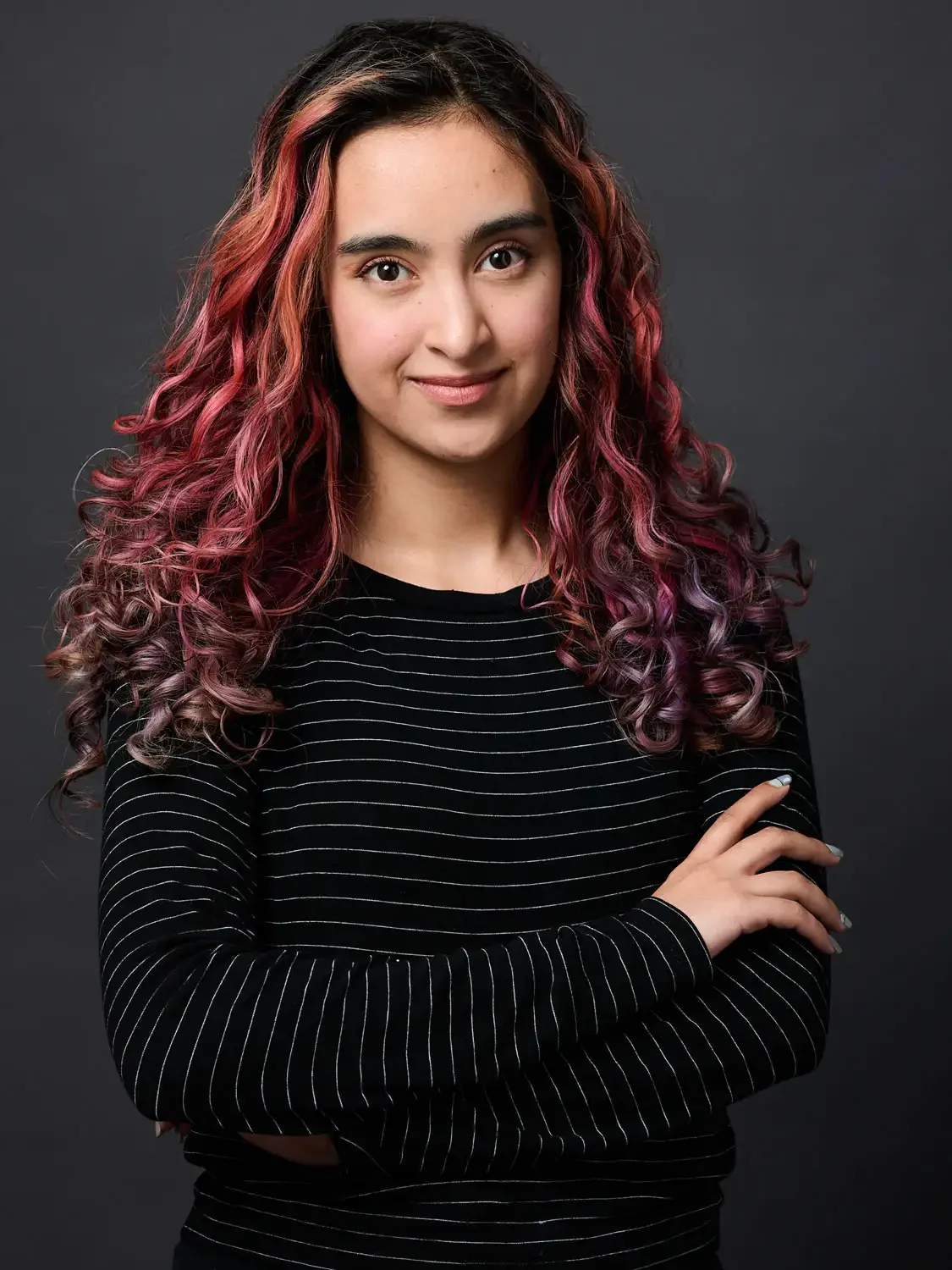 Professional headshot of a young actress with colorful, curly pink and purple hair, wearing a black and white striped shirt.