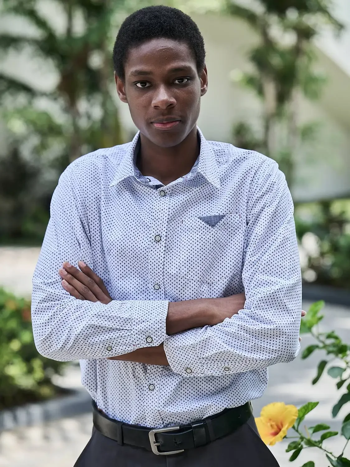 Outdoor headshot of a young man with dark hair and dark skin, in front of a blurred background of trees and flowers.