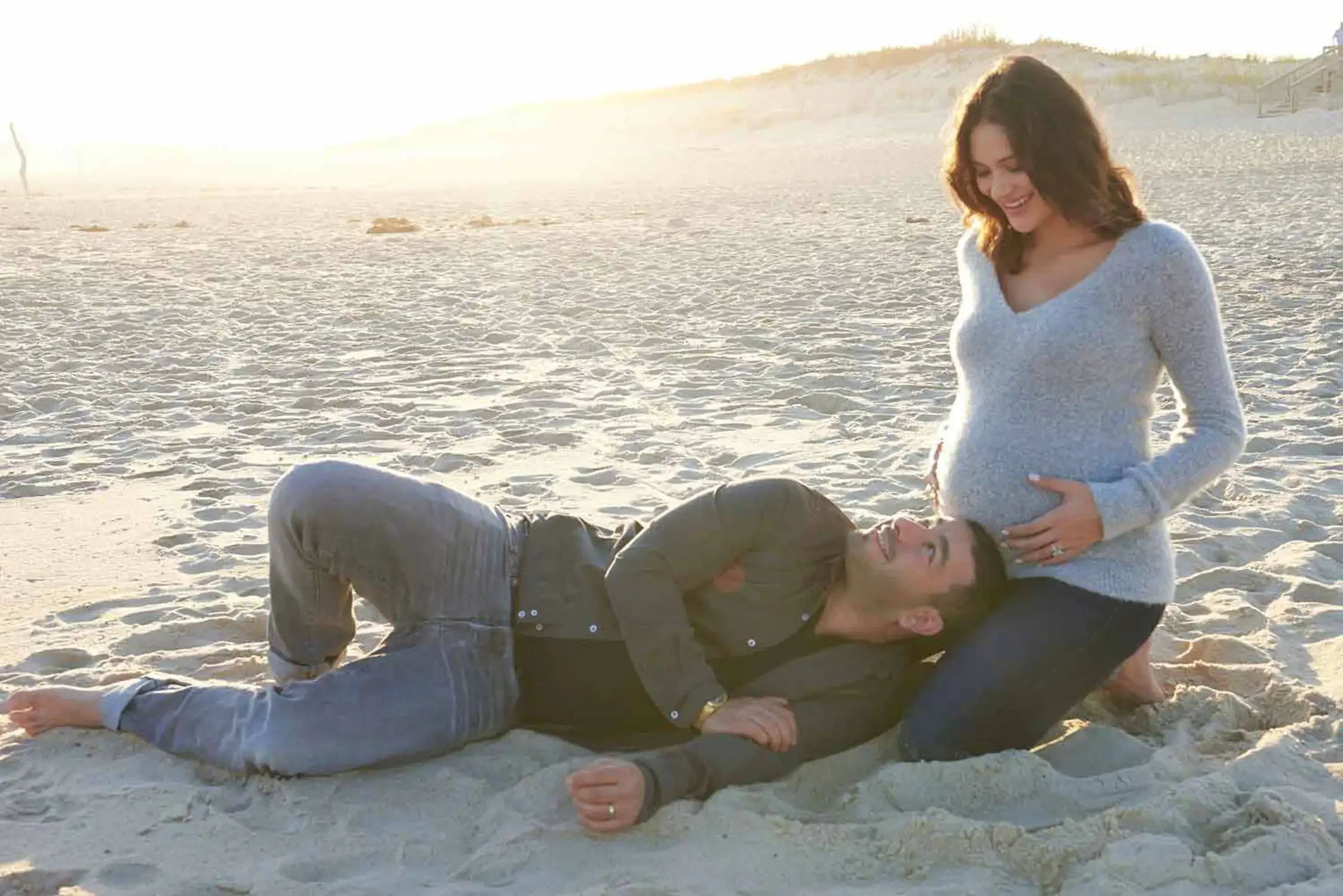 A woman laughs while looking at a man lying on the sand at the beach. The woman has shoulder-length brown hair is pregnant. The man has dark hair and is dressed in a dark jacket and pants. The scene is brightly lit by sunlight with dunes in the Hampt