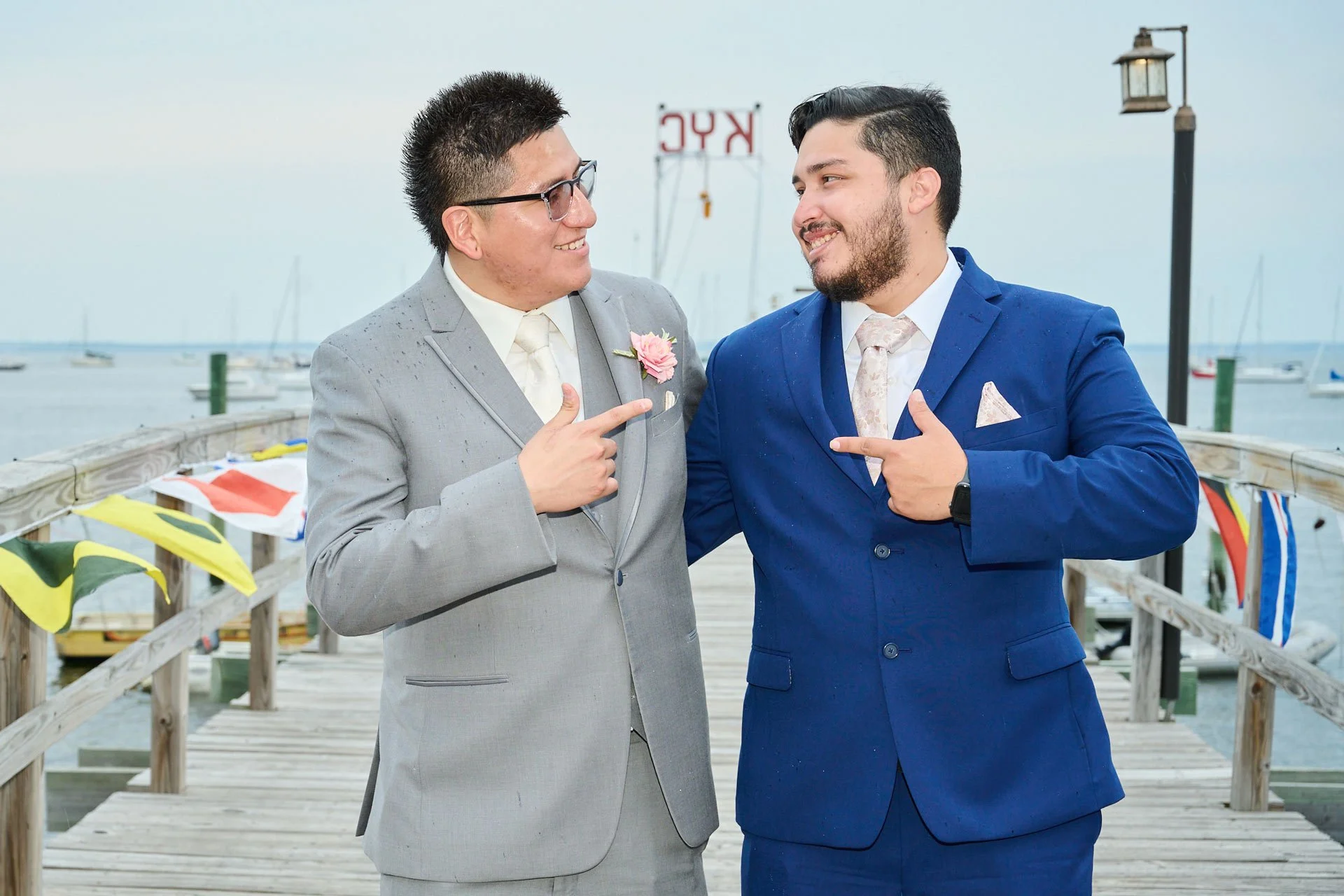 Two men in suits pointing at each other and smiling on a dock with boats and a sign in the background.