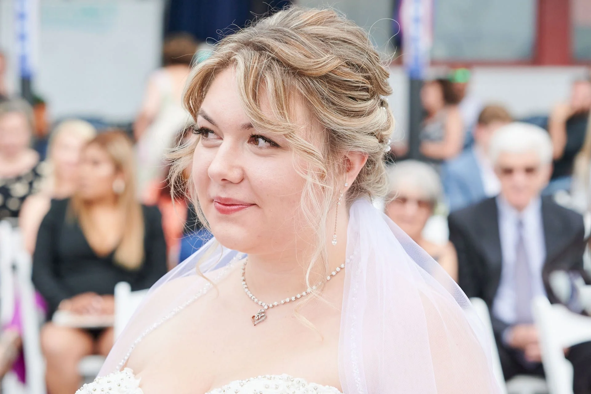 A bride with blonde hair styled in soft curls, wearing pearl jewelry and a white wedding dress, at her wedding ceremony with guests blurred in the background.