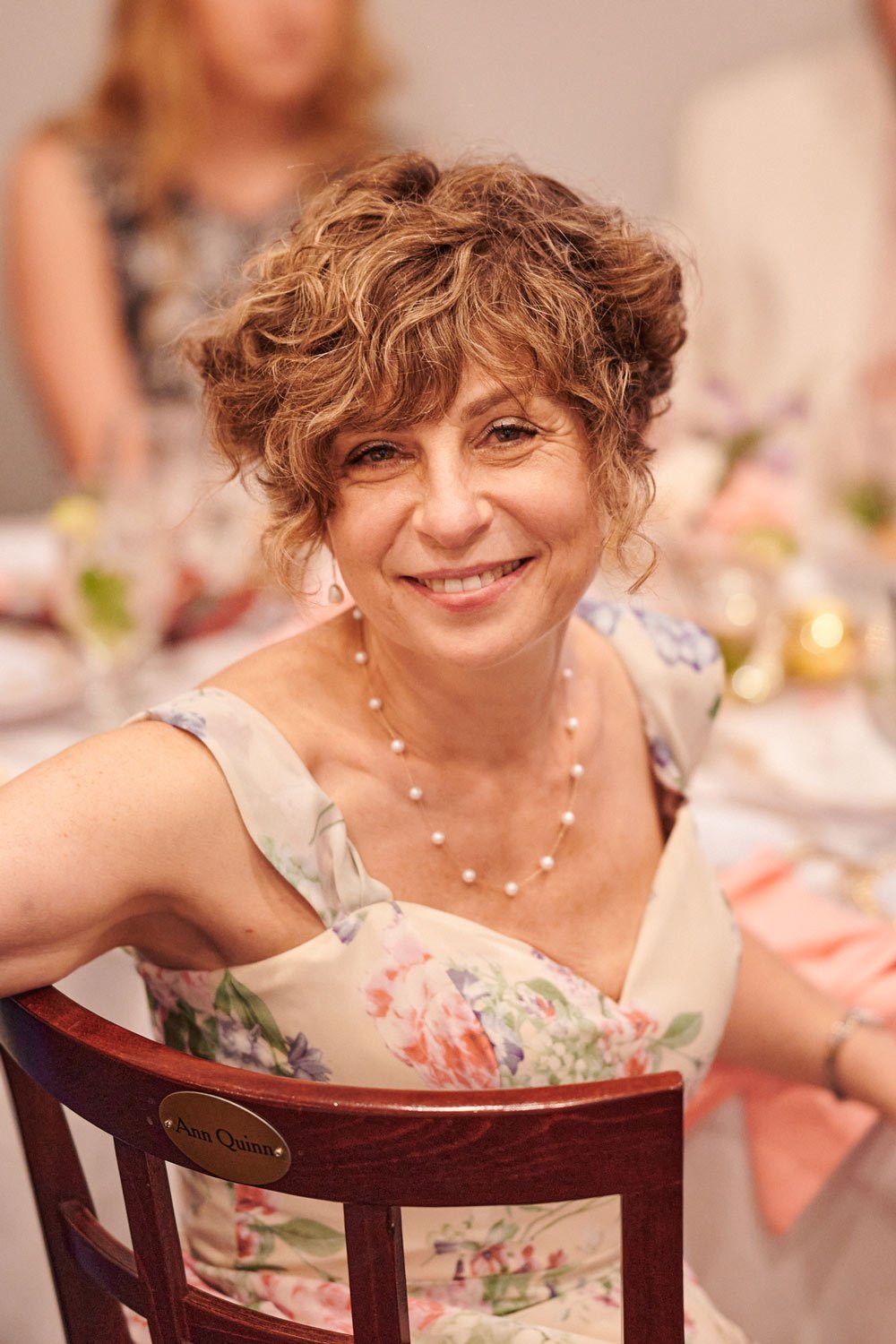 A smiling woman with curly short hair, wearing a floral dress, pearl necklace, and earrings, sitting at a decorated table during a celebration.