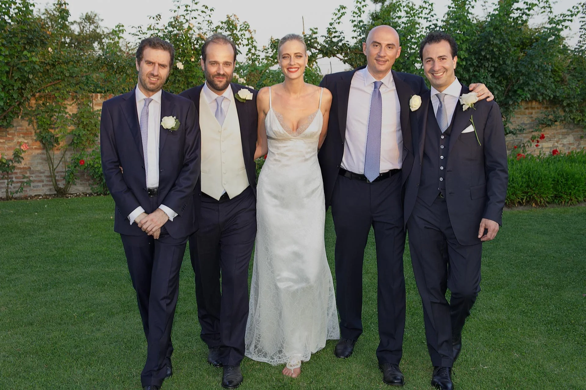 Group of six people, including one woman in a wedding dress and five men in suits, standing outdoors on a grass lawn with trees and a brick wall in the background.