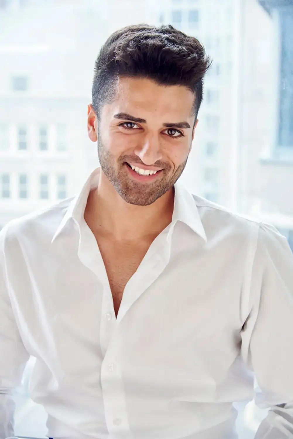 A smiling man with dark hair and a beard, wearing a white shirt, in front of large windows photographes with a cityscape background.
