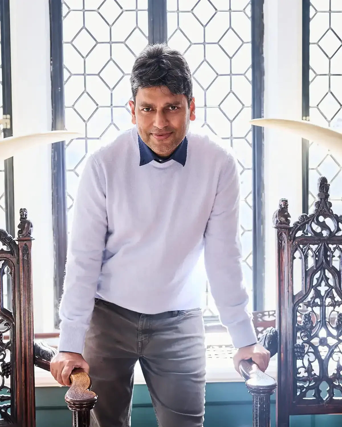 Headshot man leaning forward on a wooden railing in front of a decorative window, indoors, wearing a white sweater over a dark collared shirt, smiling at the camera.