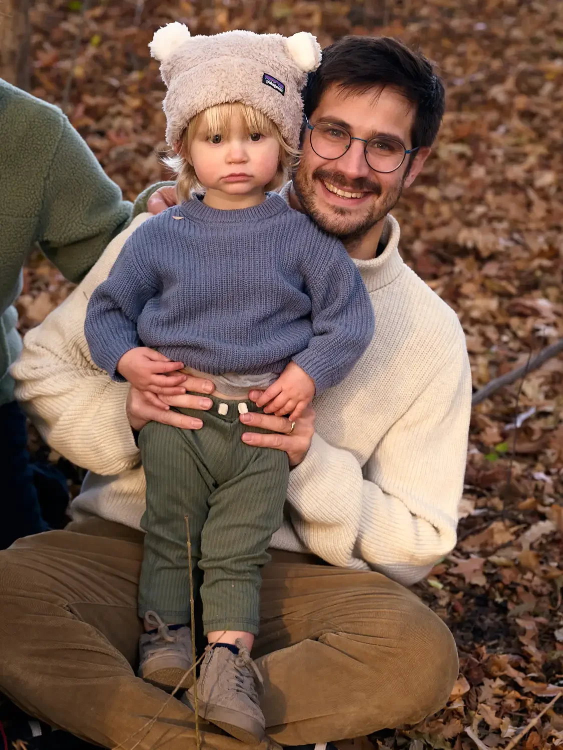 Family photo of a  man with glasses and a beard, smiling, holding a young child with blonde hair, wearing a bear hat and a blue sweater, in an outdoor setting with fallen leaves.