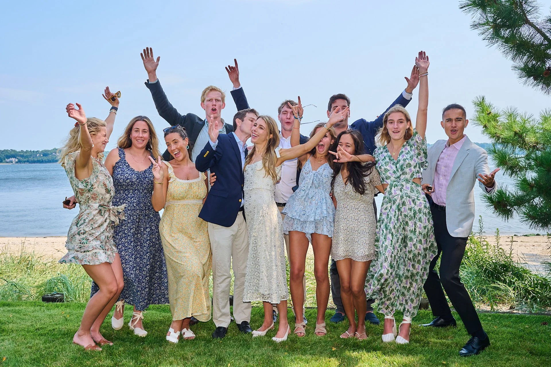Group of young adults celebrating outdoors on a grassy area near a lake, dressed in summer clothing, with some smiling, dancing, and making gestures; a tree on the right side and a blue sky overhead.