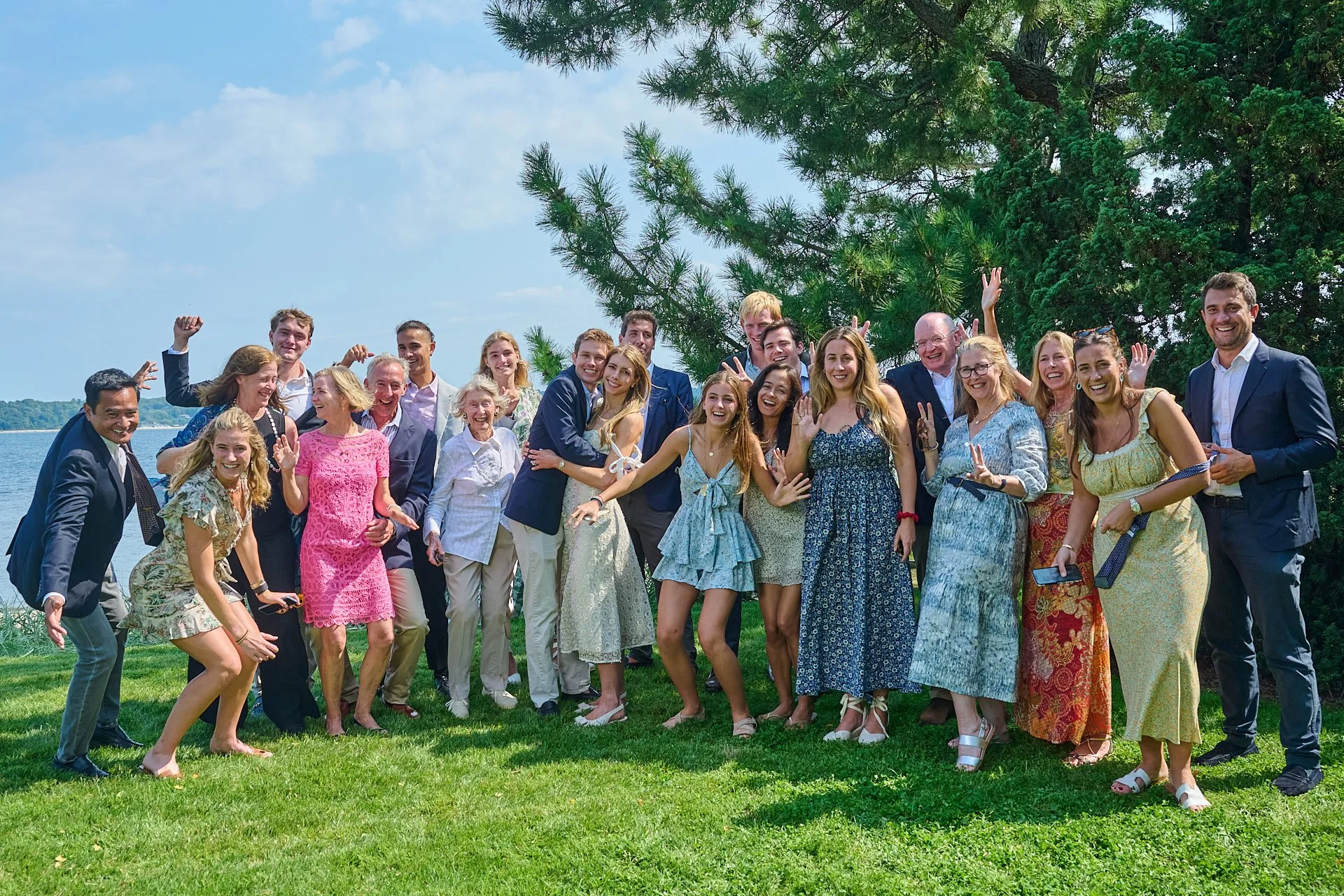 Group of smiling people celebrating outdoors by a lake with trees and a blue sky