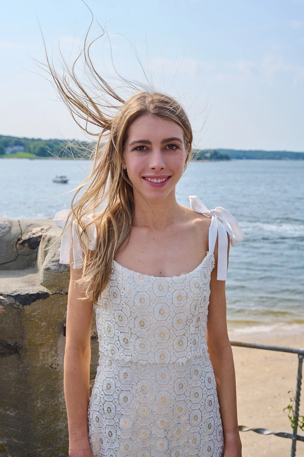 A young woman with long blonde hair stands outdoors near a body of water, wearing a white dress with floral cutout details and tied shoulder bows, smiling at the camera with wind blowing her hair.
