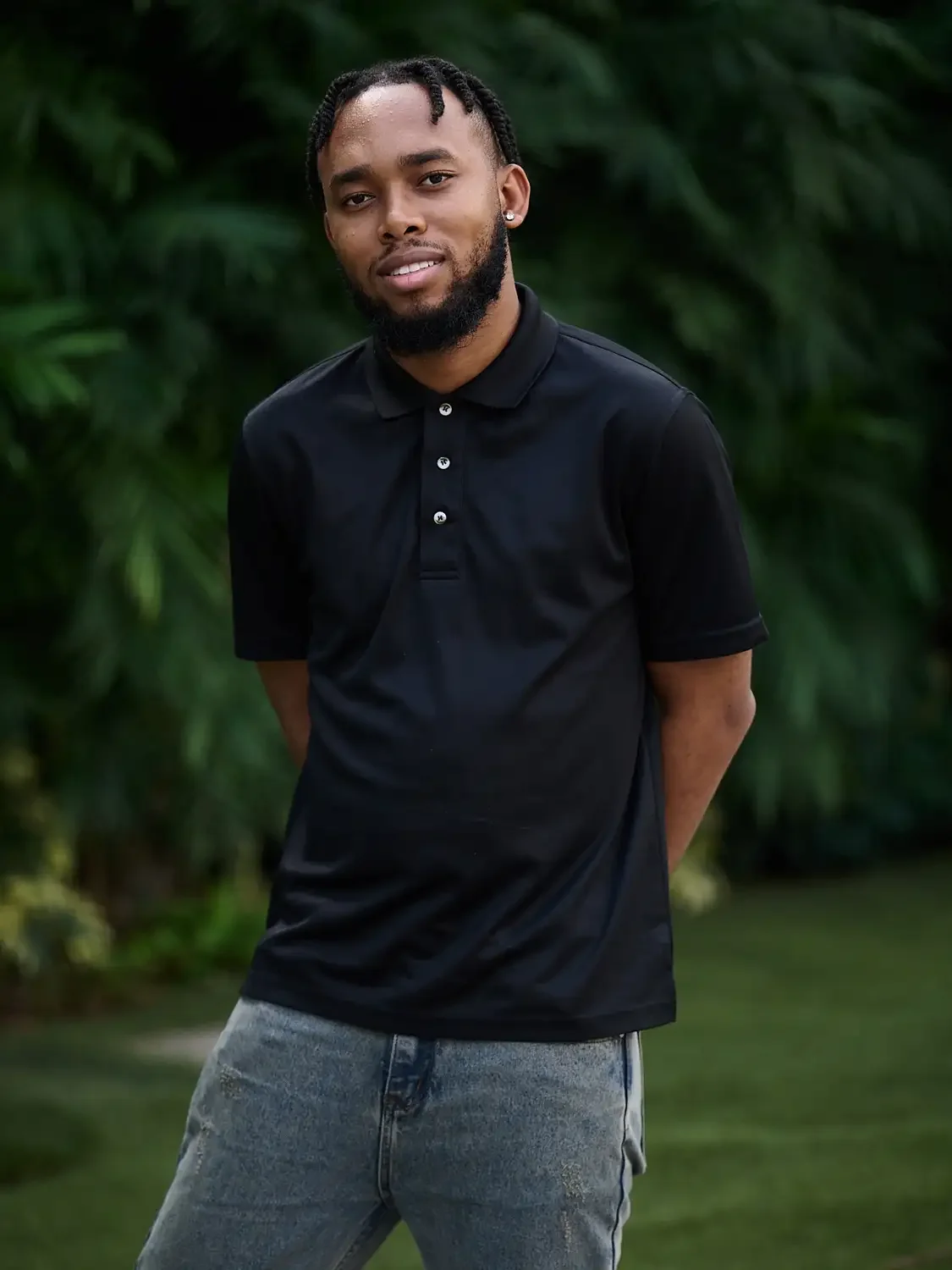 Professional photoshoot of a young man with a beard and braided hair, wearing a black polo shirt and jeans, standing outdoors with greenery in the background.