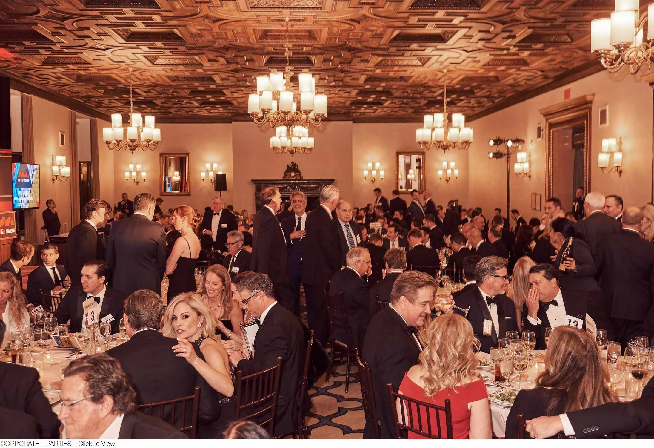 Formal banquet with guests in tuxedos and evening gowns seated at round tables, with some people standing and socializing, in an elegant decorated room with chandeliers and wall sconces.