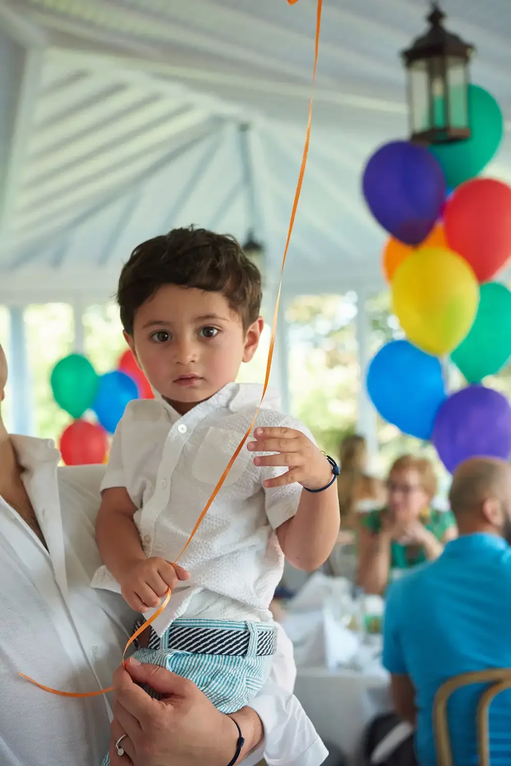 A young boy holding a balloon at a birthday party with colorful balloons and guests in the background.
