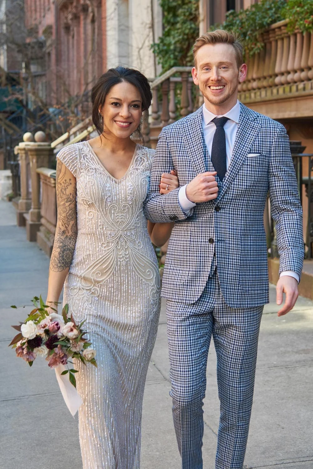 A smiling woman in a white, beaded wedding dress holding a bouquet, walking arm-in-arm with a man in a blue checkered suit and tie, on a city sidewalk during daylight.