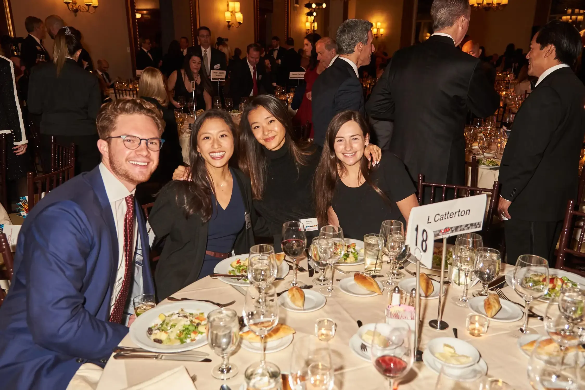 People gathered at a formal dinner event in an elegant banquet hall, with table settings, plates of salad, bread rolls, glasses of wine.