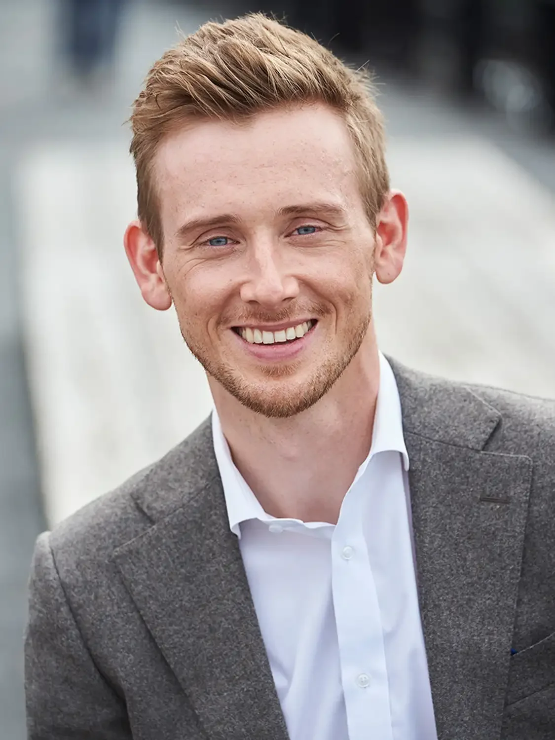 A young man with red hair, blue eyes, and light skin, wearing a gray blazer and white shirt, smiling photographed outdoors.