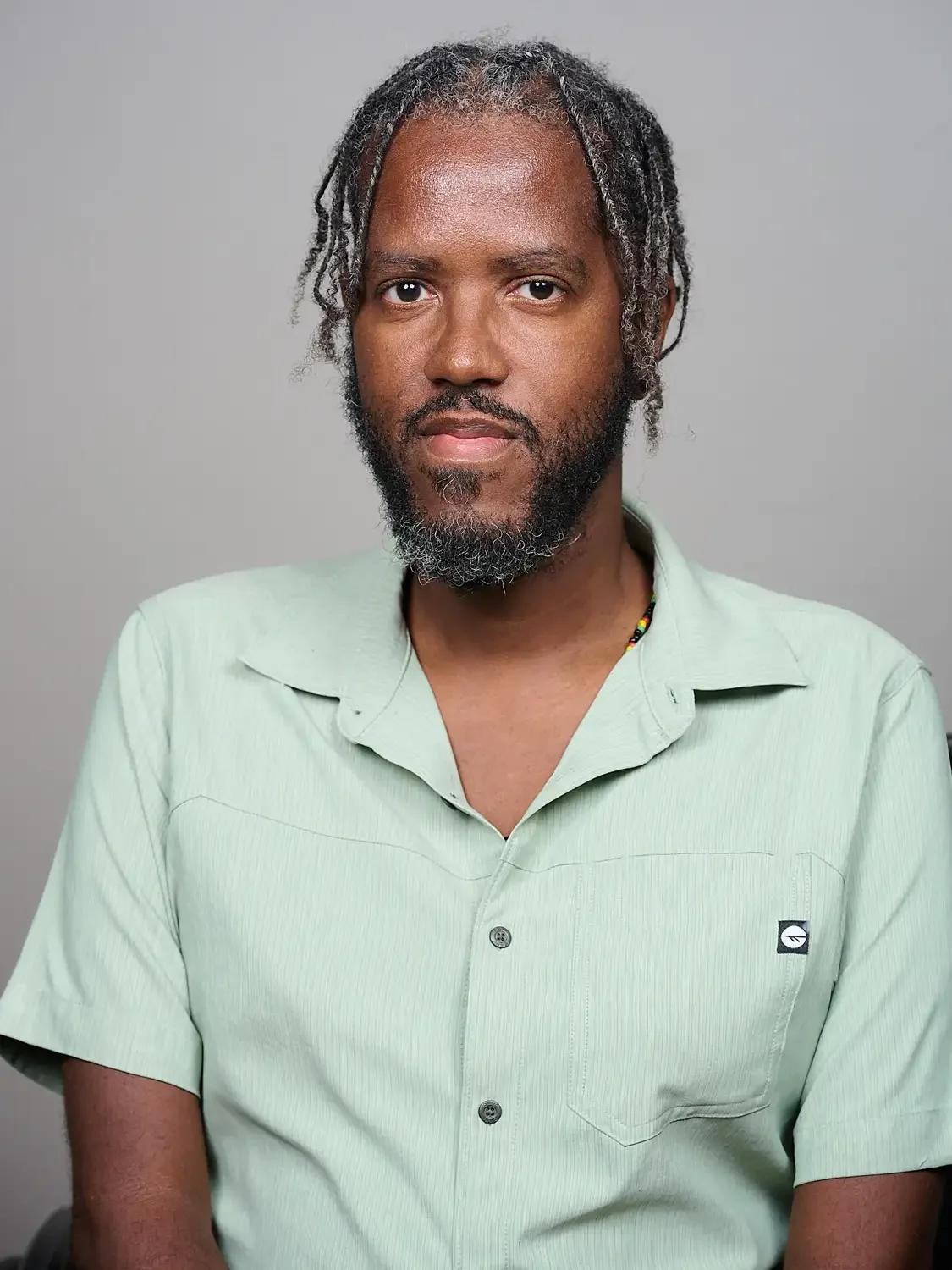 Professional portrait of a man with dreadlocks, beard, and mustache wearing a light green button-up shirt with a small logo on the pocket.
