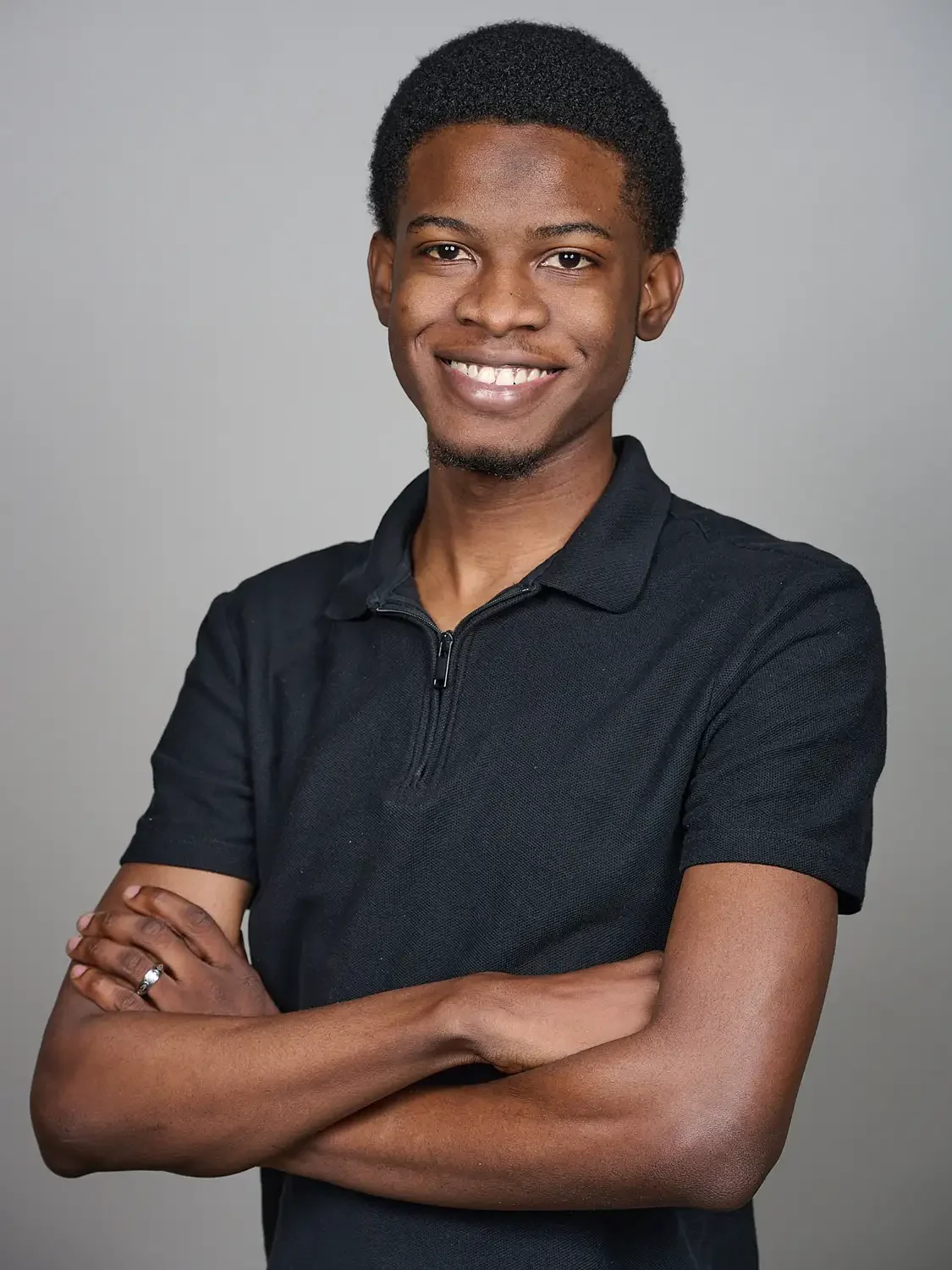 A young man with short, curly black hair, smiling, arms crossed, wearing a black polo shirt. Photographed in a professional photo studio.