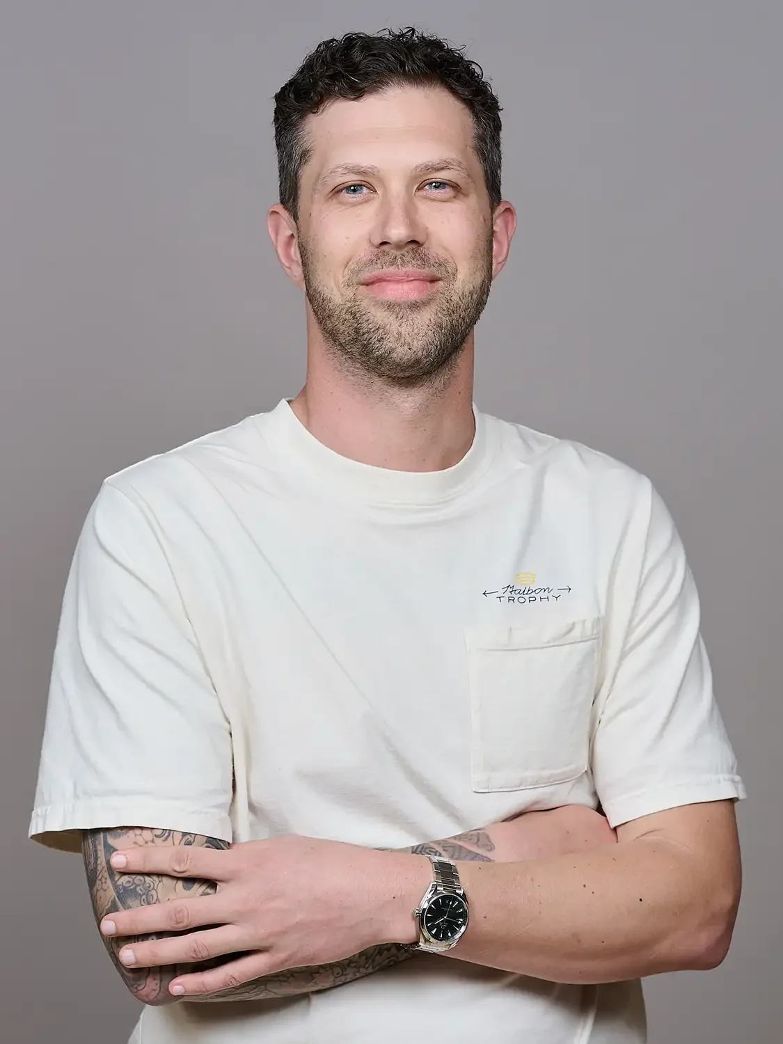 A man with short dark hair, a beard, and tattoos on his arms, wears a white T-shirt with a small logo and a watch smiling and confident attitude.