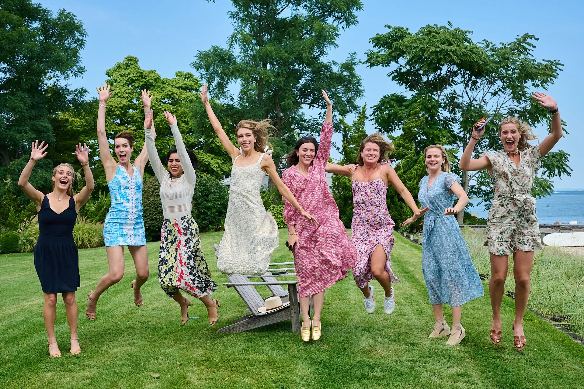 Group of eight women in colorful summer dresses jumping and celebrating outdoors on green grass near trees and water.