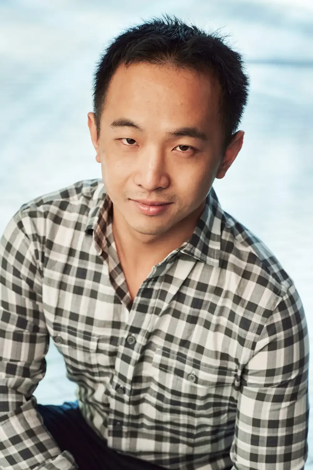 Portrait of a young man with short black hair, wearing a black and white checkered shirt, smiling slightly while looking into the camera.