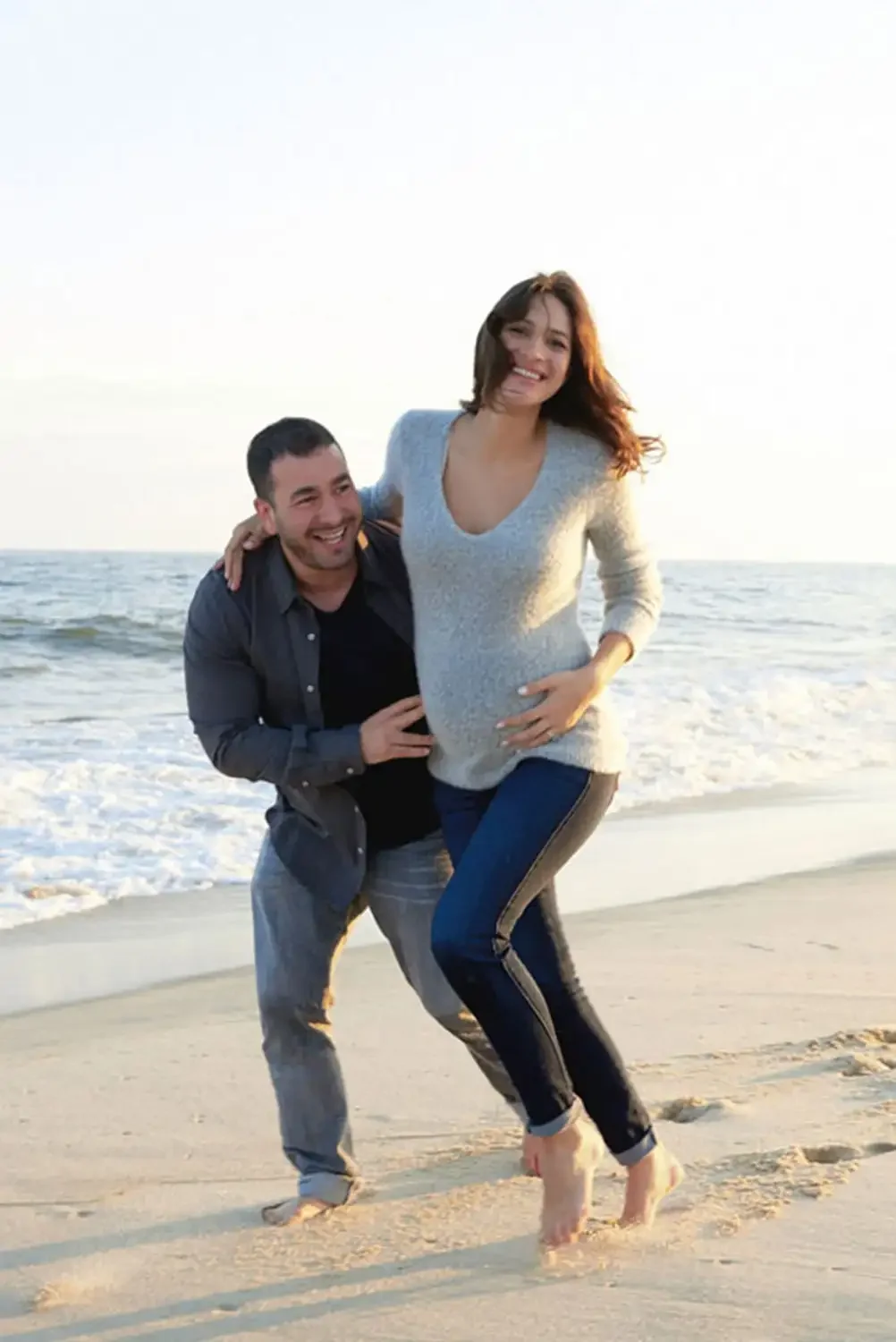 A couple on the beach, the woman is pregnant and wearing a light-colored sweater and jeans, while the man is kneeling next to her, wearing a dark shirt and light jeans, both smiling and enjoying their time in the Hamptons