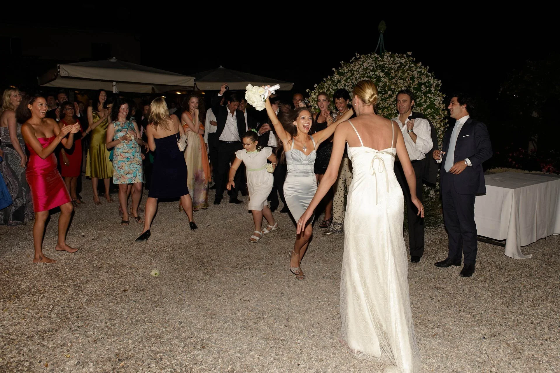 A bride in a white dress is dancing and throwing a bouquet at a wedding reception at night. Guests are celebrating around her, some clapping and others taking pictures. There are large floral arrangements and umbrellas in the background.